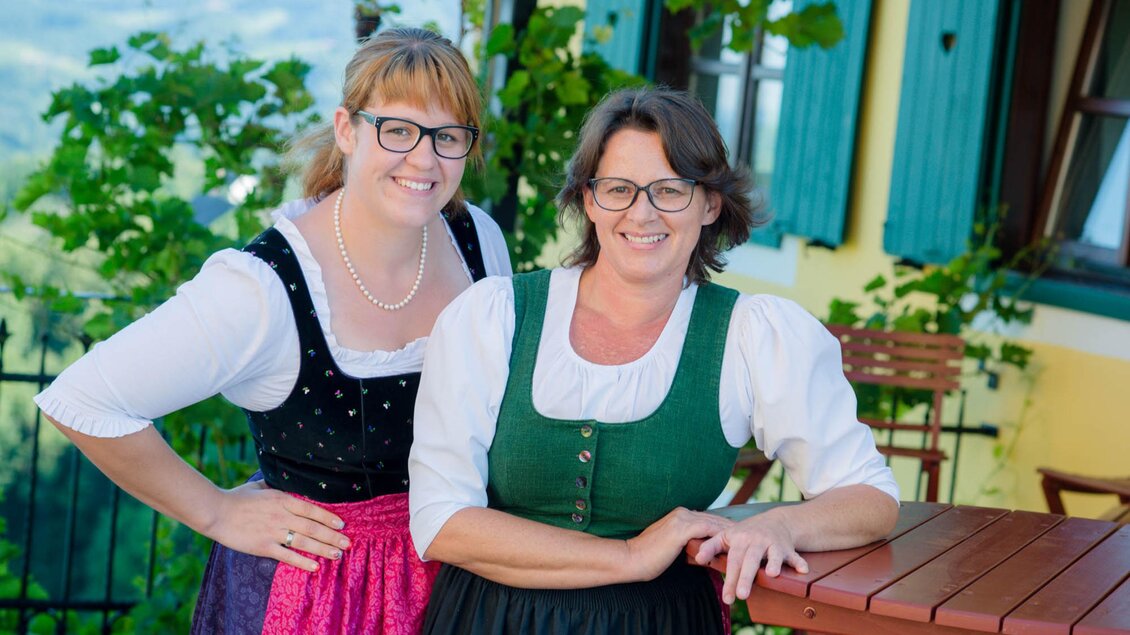 Zwei Frauen in traditioneller Tracht stehen lächelnd vor einem Haus mit grünen Fensterläden. Um sie herum sind Pflanzen und ein Tisch im Freien. | © Familie Repolusk | Weingut Repolusk