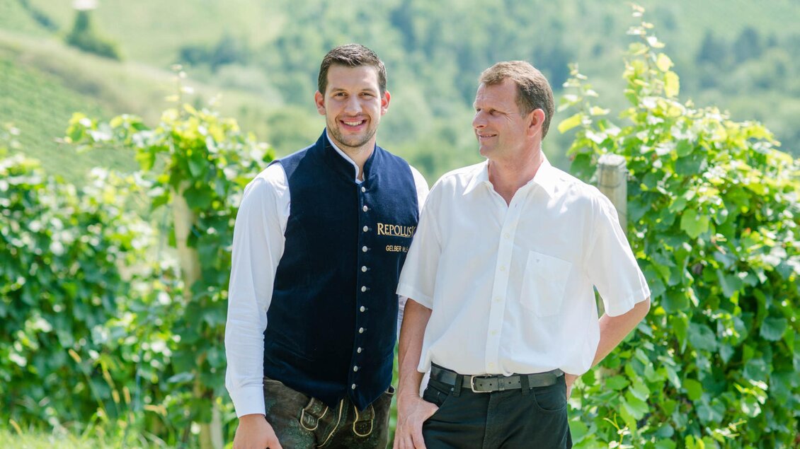 Zwei Männer stehen in einem Weinberg. Im Hintergrund sind grüne Weinreben und sanfte Hügel zu sehen. | © Familie Repolusk | Weingut Repolusk