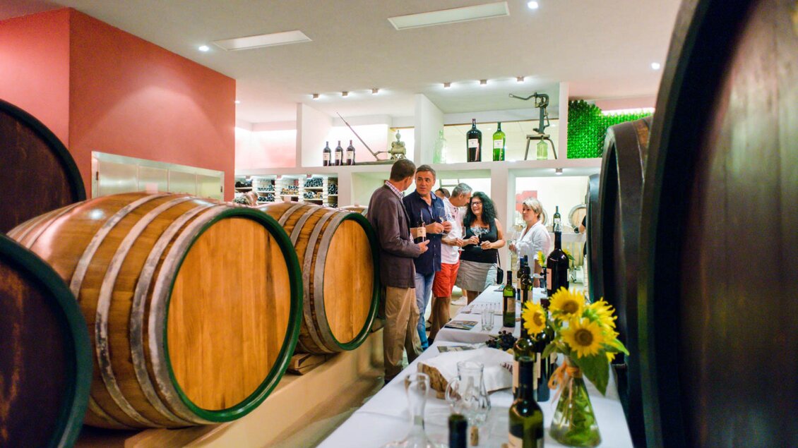 Eine Weinverkostung in einem eleganten Weingut. Im Vordergrund sind große Weinfässer und eine Tafel mit Weinflaschen und Gläsern. | © Familie Repolusk | Weingut Repolusk