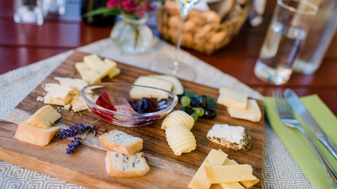Ein Holzbrett mit verschiedenen Käsesorten, begleitet von Marmelade und Trauben. Im Hintergrund sind Blumen und Gläser sichtbar. | © Familie Repolusk | Weingut Repolusk