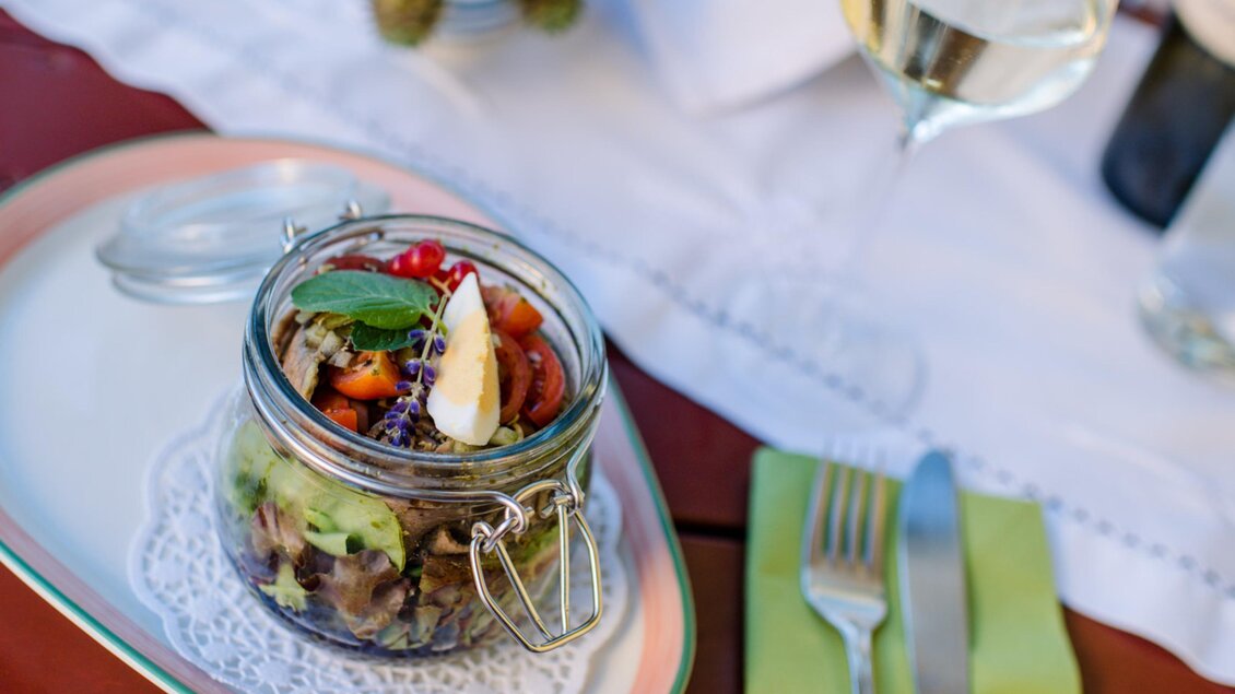 Ein bunter Salat in einem Glas mit frischem Gemüse und Kräutern. Im Hintergrund steht ein Glas Weißwein auf einem gedeckten Tisch. | © Familie Repolusk | Weingut Repolusk