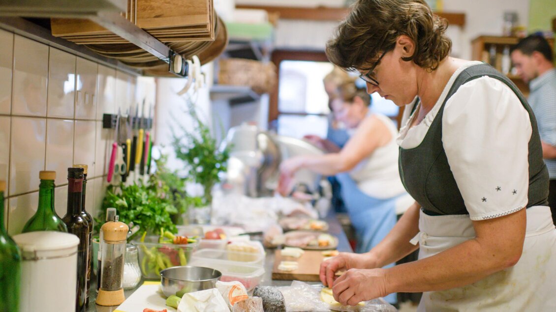 Eine Person in traditioneller Kleidung bereitet Lebensmittel in einer Küche vor. Im Hintergrund sind weitere Personen und verschiedene Kochutensilien zu sehen. | © Familie Repolusk | Weingut Repolusk