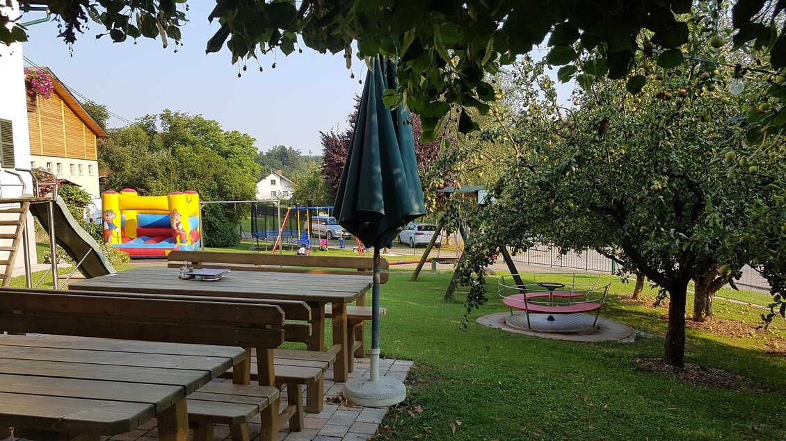 Ein einladender Garten mit Holztischen und Stühlen. Im Hintergrund sind Spielgeräte und ein großer Baum zu sehen. | © Buschenschank Safner