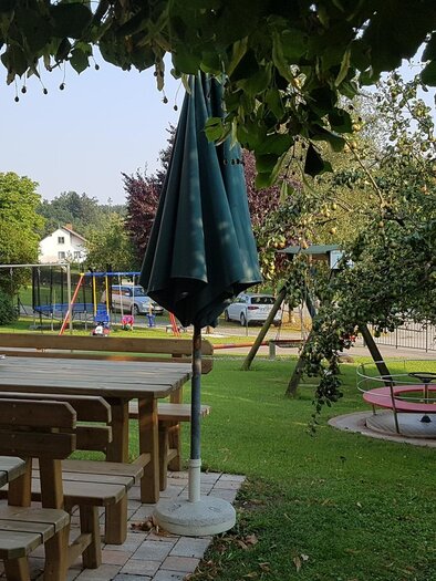 Ein einladender Garten mit Holztischen und Stühlen. Im Hintergrund sind Spielgeräte und ein großer Baum zu sehen. | © Buschenschank Safner