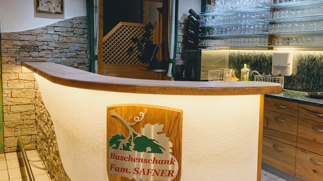 Ein einladender Barbereich mit Holzdetails und einem Schild der Familie Safner. Die Wände sind mit Stein dekoriert, und zahlreiche Gläser sind ordentlich aufgereiht. | © Buschenschank Safner