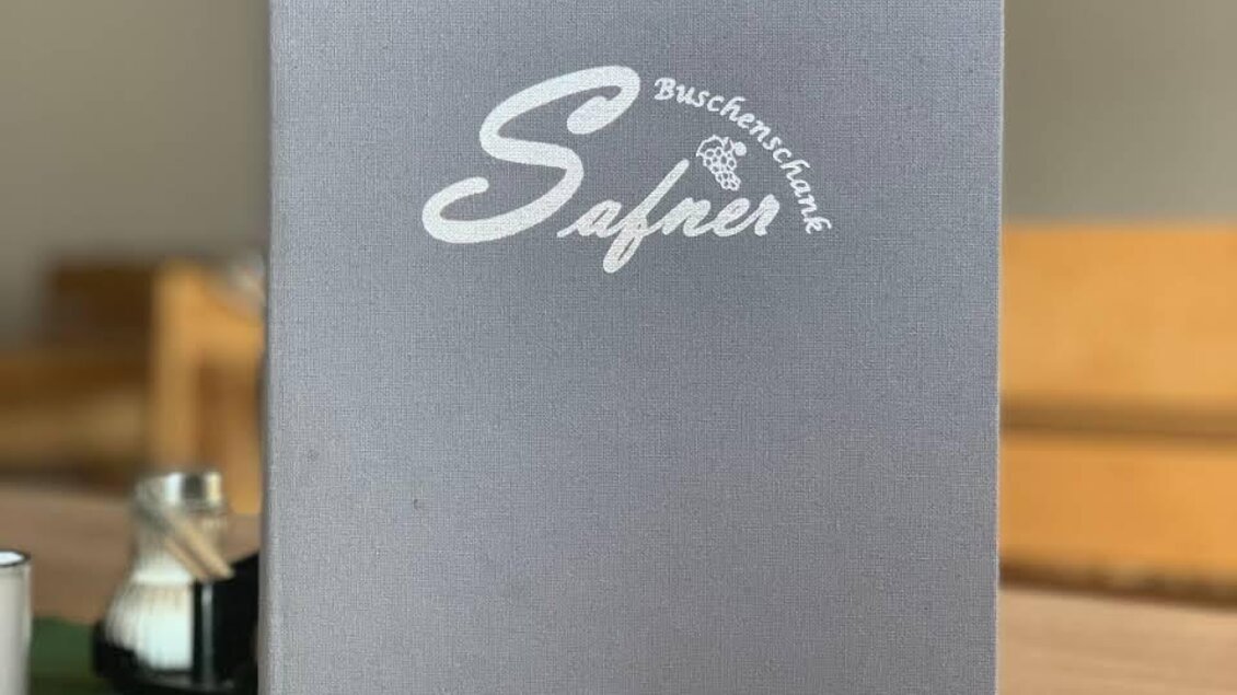 Ein graues Menübuch mit dem Schriftzug "Safner" und der Aufschrift "Buschenschank". Im Hintergrund sind eine Kaffeekanne und ein Tisch zu sehen. | © Buschenschank Safner