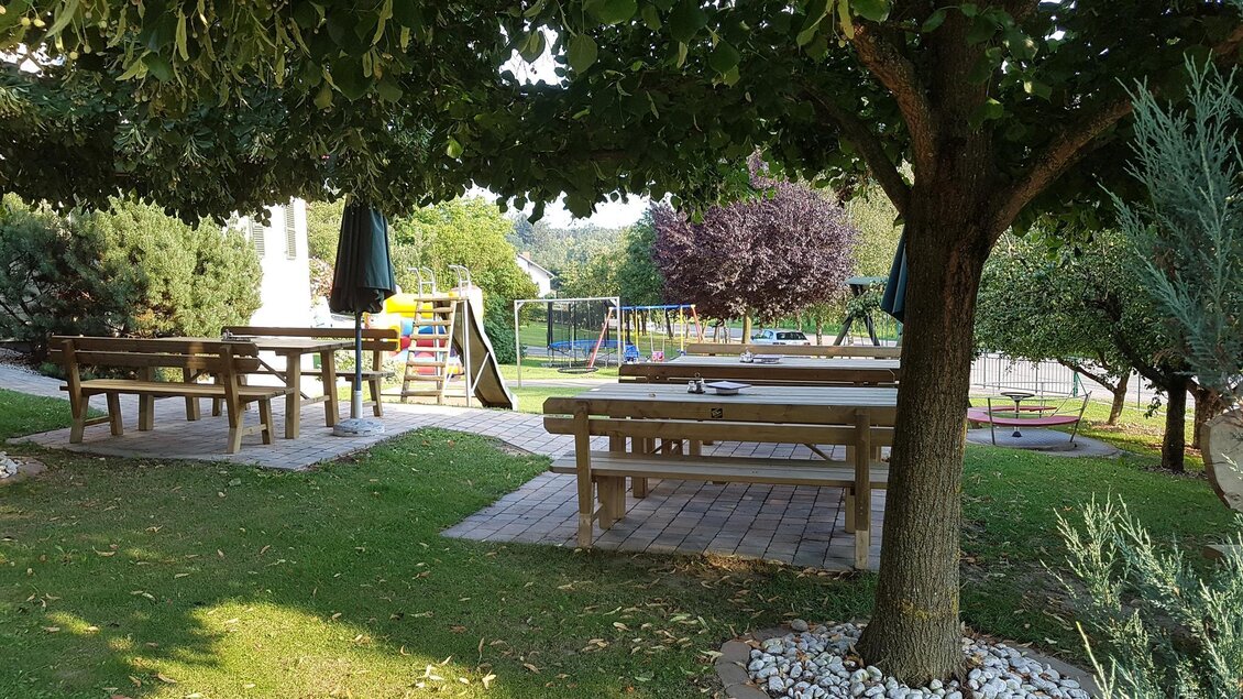 Ein schöner Garten mit Holztischen und Bänken unter Bäumen. Im Hintergrund ist ein Spielplatz sichtbar. | © Buschenschank Safner