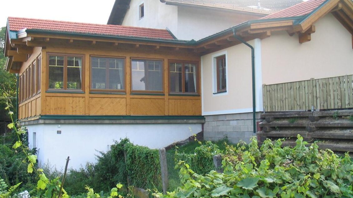 Ein modernes Haus mit einem überdachten Anbau aus Holz. Der Garten ist grün und von Pflanzen umgeben. | © Buschenschank Schachner