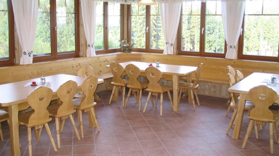 Ein gemütlicher Gastraum mit Holztischen und -stühlen. Große Fenster lassen viel Tageslicht herein und bieten einen Blick auf die Natur. | © Buschenschank Schachner