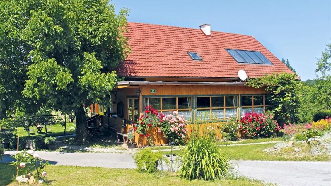 Ein gemütliches Holzhaus mit einem roten Dach und einem großen Garten. Blumen und Pflanzen umgeben das Haus, und es gibt einen blauen Himmel. | ©  Buschenschank Schulter