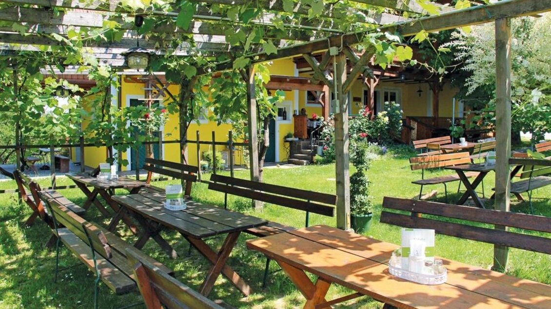 Ein einladender Gartenbereich mit Holztischen und -bänken unter einer Pergola. Im Hintergrund sind grüne Pflanzen und ein gelbes Gebäude zu sehen. | ©  Buschenschank Schulter