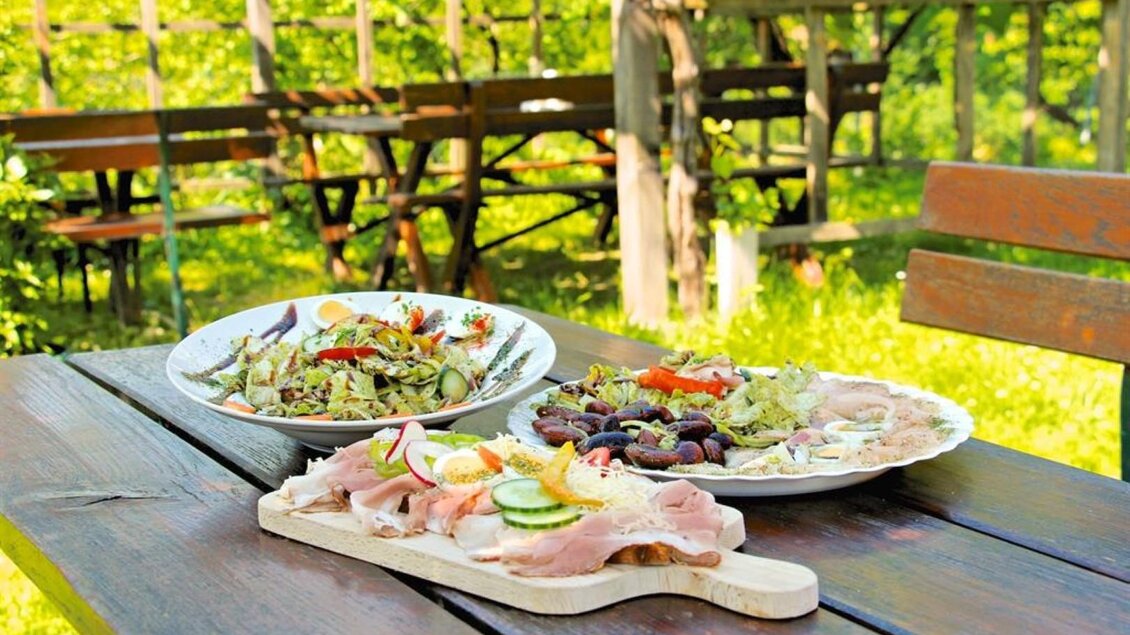 Eine gemütliche Tischdekoration im Freien mit zwei Tellern. Der erste Teller enthält Salat, der zweite eine Auswahl an Wurst und Aufschnitt. | ©  Buschenschank Schulter