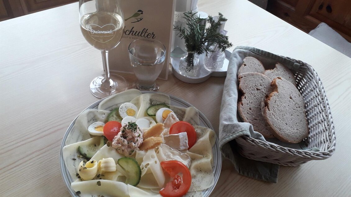 Ein Teller mit frischem Salat, Eier, und Gemüse, serviert mit Brot und einem Glas Weißwein. Im Hintergrund ist ein Tisch mit einem Menü in Aussicht. | ©  Buschenschank Schulter