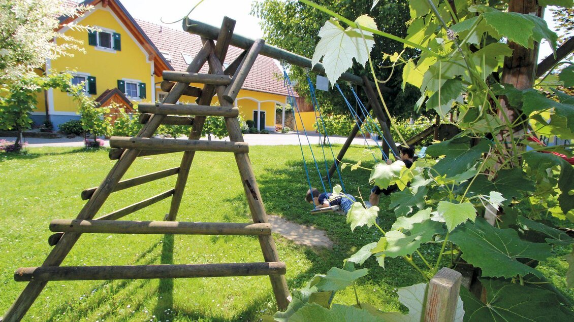 Ein Spielplatz mit einer Holzschaukel und einer Rutsche im Garten. Im Hintergrund sind gelbe Häuser und grünes Gras zu sehen. | ©  Buschenschank Schulter