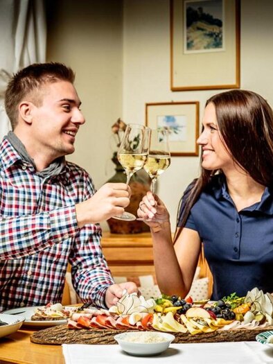 A couple raises glasses amidst a festive table decoration. Various dishes and snacks are on the table. | © Stephan Friesinger