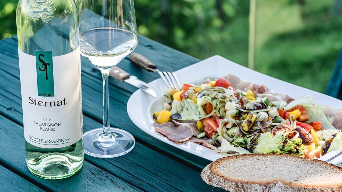 Ein leckeres Gericht mit frischem Salat, Tomaten und Schinken auf einem Teller. Dazu ein Glas Sauvignon Blanc und eine Flasche Wein auf einem Holztisch. | © Familie Sternat | Weingut Sternat