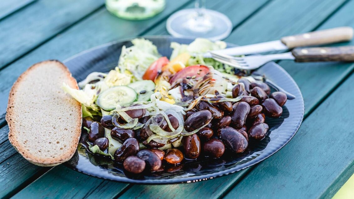 Ein Teller mit schwarzen Bohnen, frischem Gemüse und einer Scheibe Brot. Im Hintergrund ist ein Glas Wein zu sehen. | © Familie Sternat | Weingut Sternat