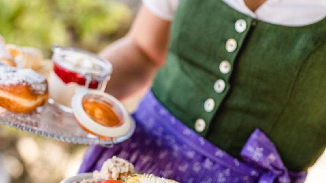 Eine Person in traditioneller Tracht hält zwei Teller mit Speisen. Auf einem Teller sind Fleisch, Gemüse und auf dem anderen Teller Desserts. | © Familie Sternat | Weingut Sternat