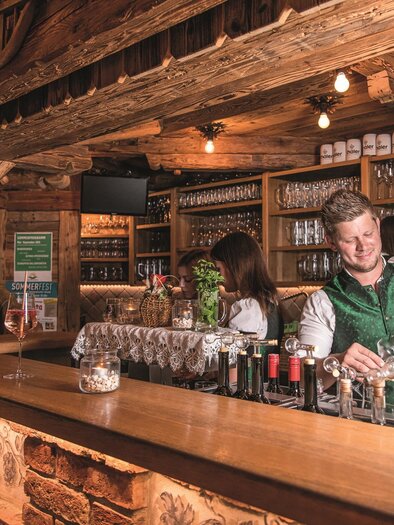 A cozy bar with wooden decor and many glasses in the background. A bartender serves drinks and creates an inviting atmosphere. | © Michaela Pfleger
