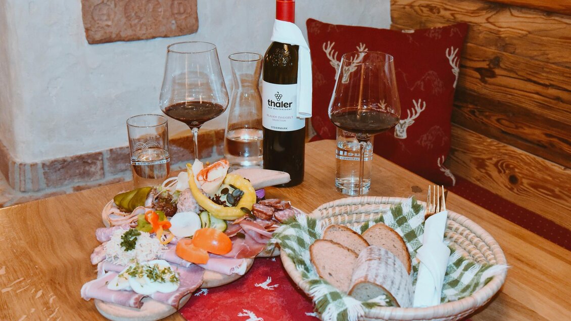 Ein rustikaler Tisch mit einer Auswahl an Aufschnitt, frischem Gemüse und Brot. Dazu stehen Gläser Wasser und Rotwein sowie eine Flasche Wein. | © Thaler Wolfgang