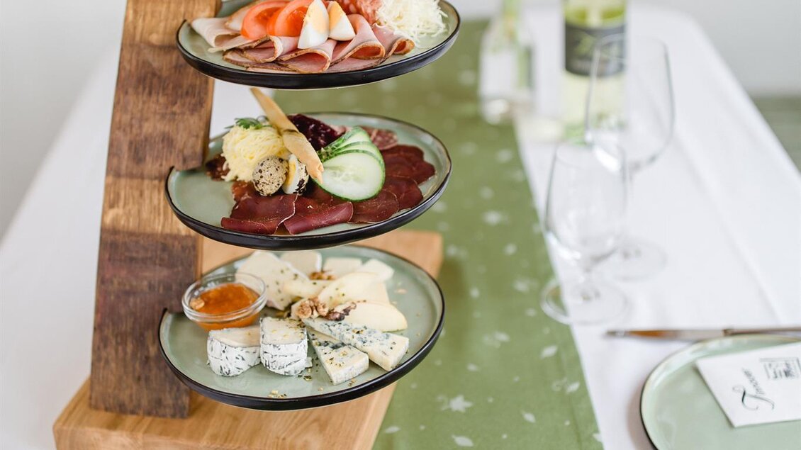 Ein dekoratives Essensturm aus verschiedenen Käsesorten, Aufschnitt und frischem Gemüse. Darunter liegt ein Salatgericht auf einem Tisch mit eleganter Tischdecke und Glaswaren. | © Tinnauer