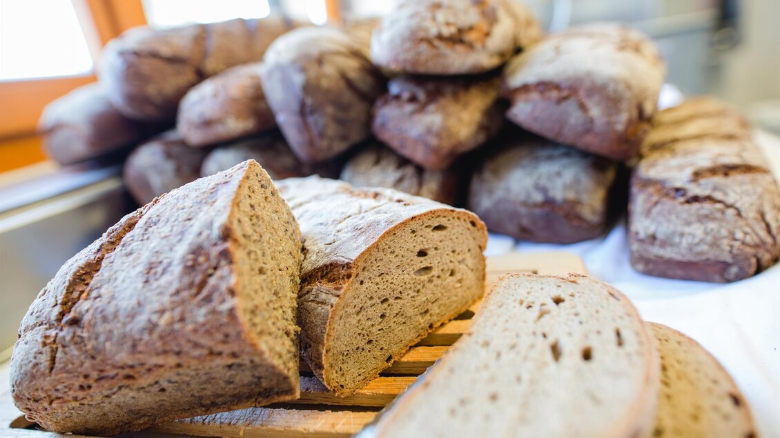 Frisches Bauernbrot, das in Scheiben geschnitten und auf einem Holzbrett präsentiert wird. Im Hintergrund sind weitere Brote gestapelt. | © Tinnauer