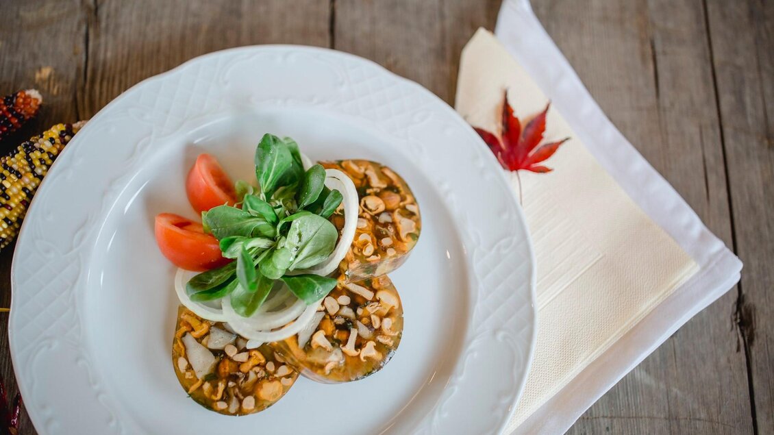 Ein schön angerichteter Teller mit einer Vorspeise aus Gemüse, Nüssen und frischen Kräutern. Dazu liegen einige Tomatenscheiben und ein Servietten auf einem rustikalen Tisch. | © Tinnauer