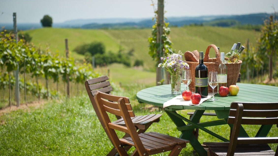 Ein gemütlicher Tisch im Freien, umgeben von Weinreben und einer schönen Landschaft. Auf dem Tisch stehen Weingläser, eine Weinflasche und frisches Obst. | © Tinnauer