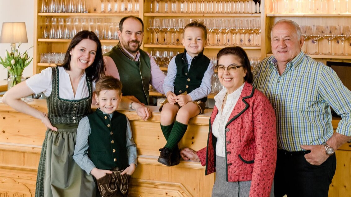 Eine Gruppe von fünf Personen in traditioneller bayerischer Tracht steht vor einer Holz-Bar. Sie lächeln freundlich und wirken einladend. | © Tinnauer