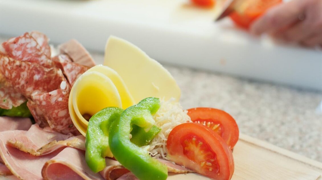 Eine Auswahl an Wurst, Käse und frischem Gemüse auf einem Holzbrett. Im Hintergrund wird Gemüse auf einem Schneidebrett geschnitten. | © Tinnauer
