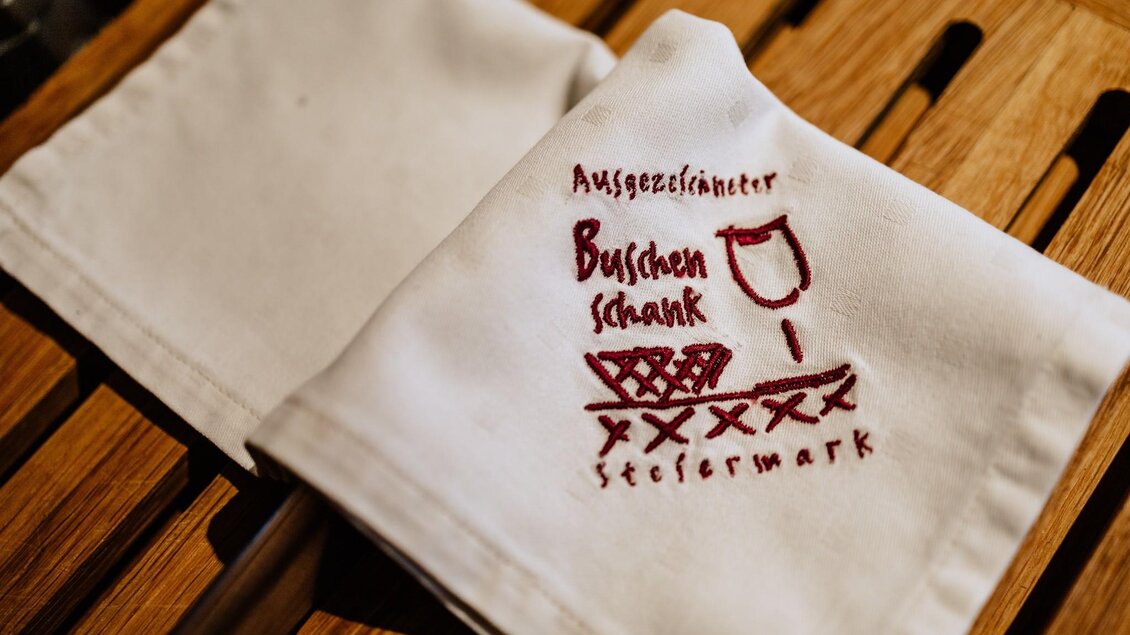 Ein weißes Serviette mit rotem Druck. Der Text zeigt "Ausgezeichneter Buschenschank Steiermark" und ein Weinglas.