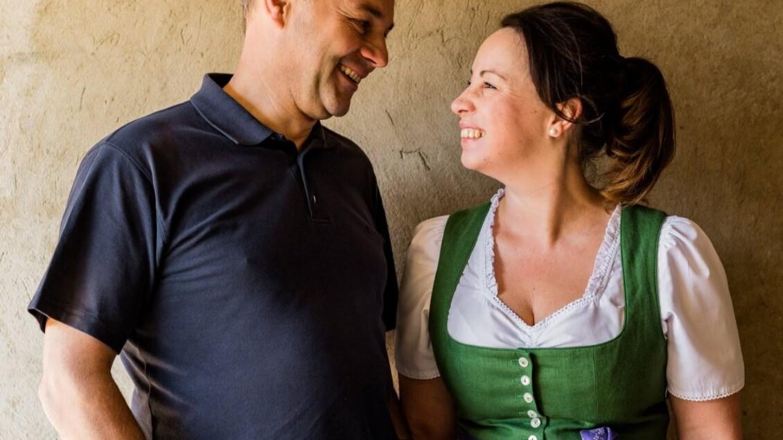 Ein Paar steht lächelnd vor einer schlichten Wand. Die Frau trägt ein traditionelles Kleid, während der Mann leger gekleidet ist.