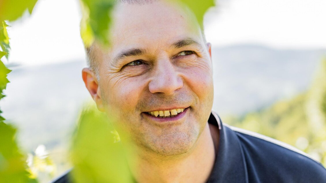 Ein lächelnder Mann steht zwischen Weinreben. Im Hintergrund sind sanfte Hügel und blauer Himmel zu sehen. | © Weingut Tschermonegg