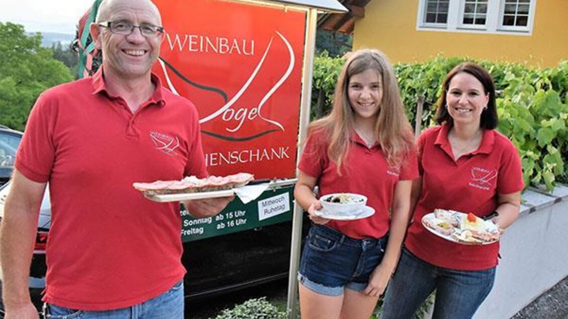 Eine Familie steht vor ihrem Weingut und serviert Essen. Alle tragen rote T-Shirts und lächeln freundlich in die Kamera.