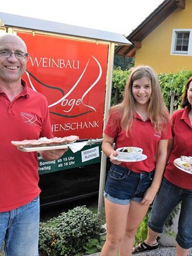 A family is standing in front of their winery and serving food. Everyone is wearing red T-shirts and smiling kindly at the camera.