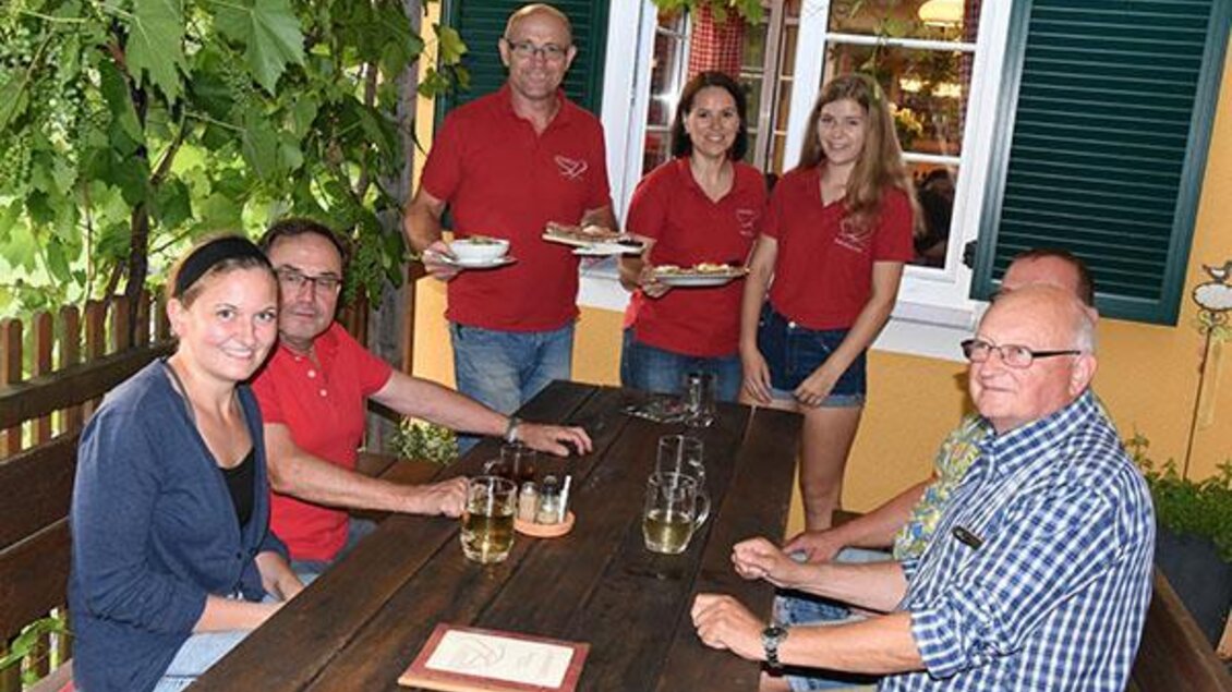 Eine Gruppe von Menschen sitzt an einem Holztisch im Freien, umgeben von Weinreben. Einige Personen in roten T-Shirts servieren Essen und Getränke.