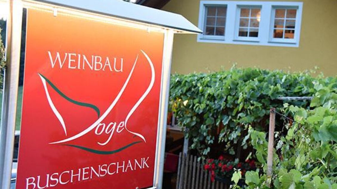 Ein rotes Schild mit der Aufschrift „Weinbau Vogel Buschenschank“. Im Hintergrund sind Fenster eines Hauses und Weinreben zu sehen.