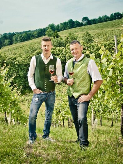 Zwei Männer stehen in einem Weinberg und halten Gläser mit Rotwein. Im Hintergrund sind grüne Weinreben und sanfte Hügel zu sehen. | © Weingut Weber