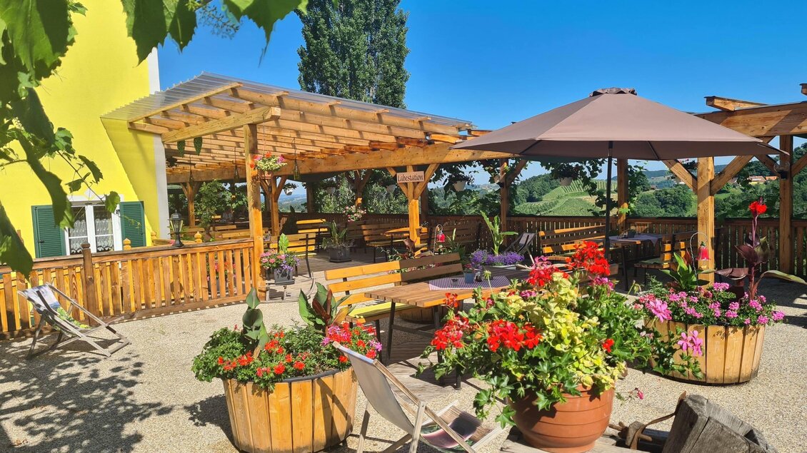 Ein einladender Außenbereich mit bunten Blumen und Holzmöbeln. Der klare Himmel und die Sonnenschirme schaffen eine gemütliche Atmosphäre. | © Weingut Wechtitsch