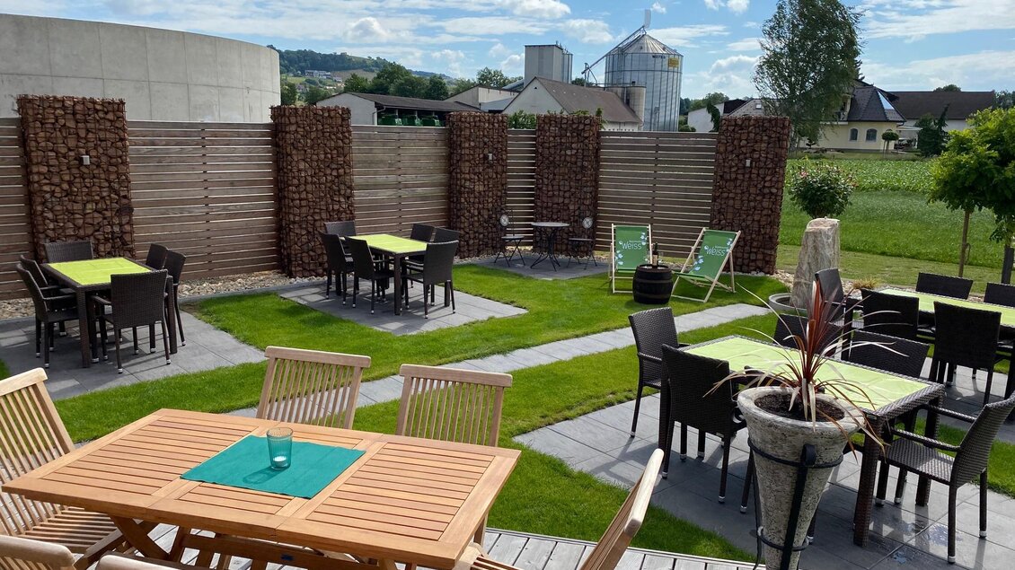 Eine gemütliche Terrasse mit Holzstühlen und Tischen im Freien. Im Hintergrund sind Wiesen und einige Gebäude zu sehen. | © BS Weiß