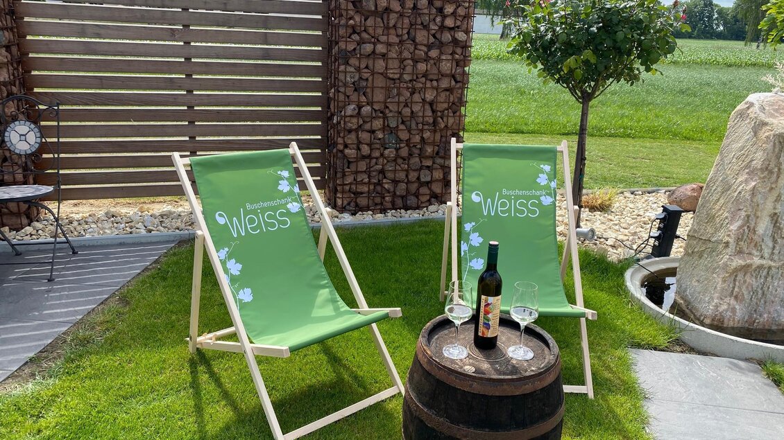 Zwei grüne Liegestühle stehen auf einer Rasenfläche neben einem Holzfass. Auf dem Tisch befindet sich eine Flasche Wein und zwei Gläser. | © BS Weiß