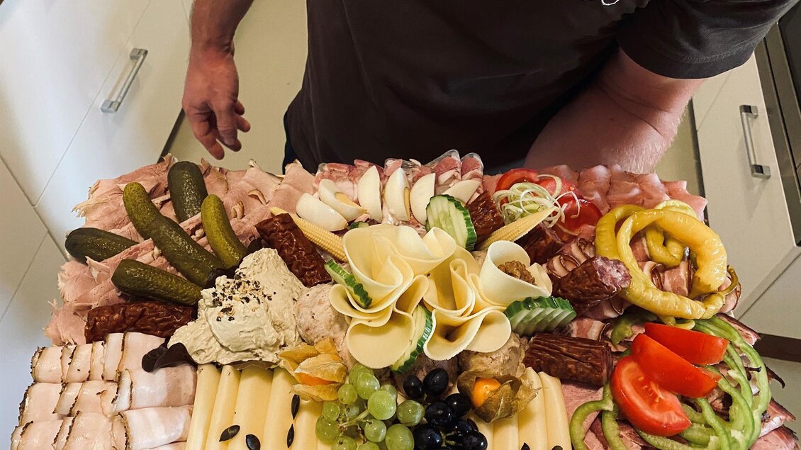 Ein reichhaltiges Wurst- und Käsebrett mit verschiedenen Aufschnitt, Käse, frischen Gemüsesorten und Trauben. Es wird von einer Person gehalten und zeigt eine bunte Auswahl an leckeren Snacks. | © BS Weiß