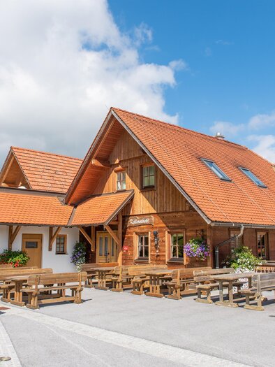 A traditional wooden house with an orange roof and a cozy terrace. The surroundings are green and inviting, ideal for a sunny weekend. | © Buschenschank Kellerstöckl