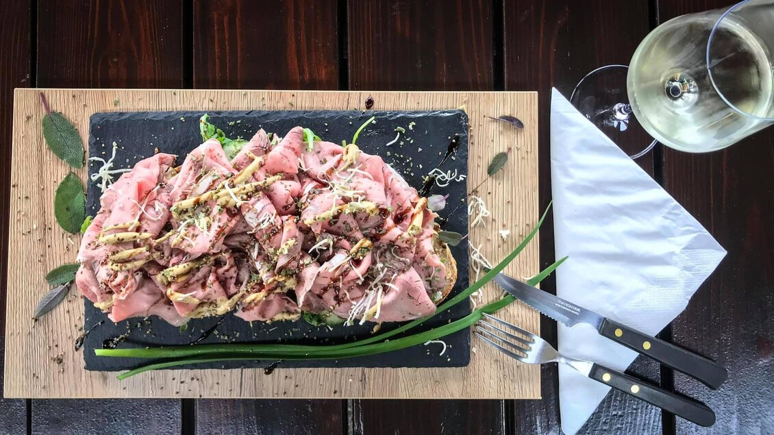 Ein ansprechend angerichteter Teller mit verschiedenen Fleischsorten und Kräutern. Dazu ein Glas Weißwein auf einem Holzuntergrund. | © Weingut Weiss-Welle | Bettina Gratzer