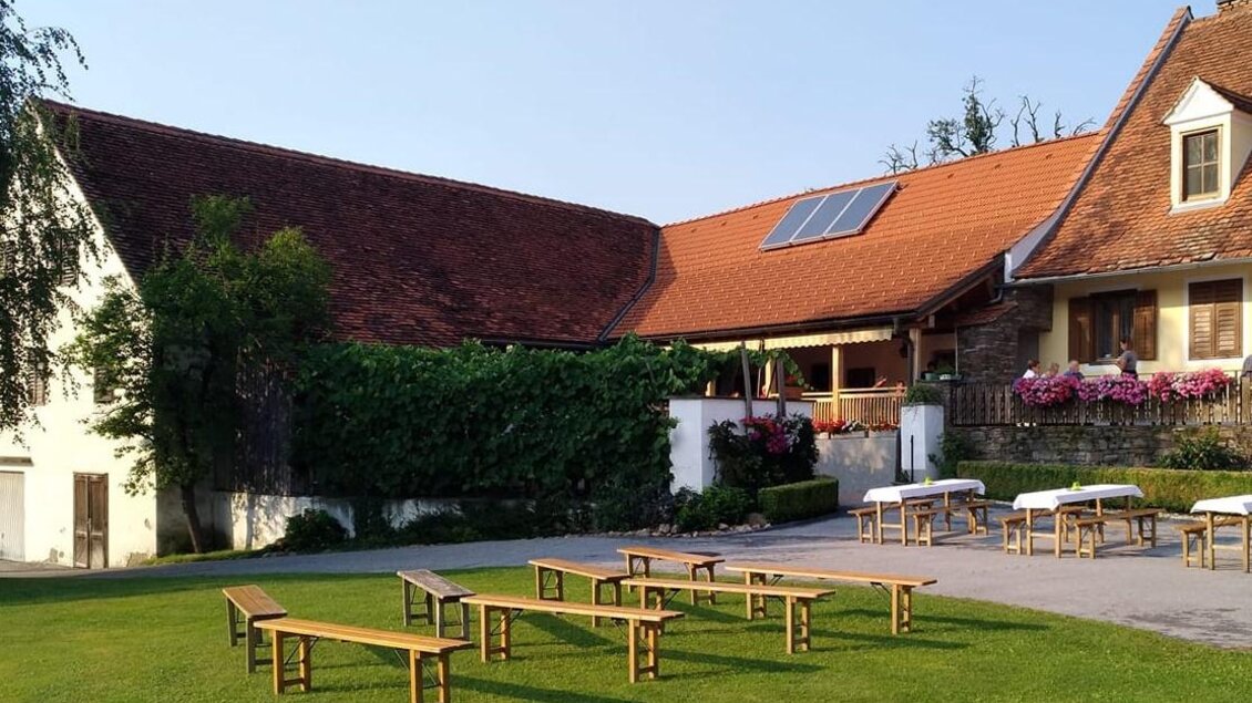 Eine ruhige Grünfläche mit Holzbänken und Tischen. Im Hintergrund ist ein traditionelles Gebäude mit einem roten Dach und Blumenschmuck zu sehen. | © Buschenschank Wiesenhofer
