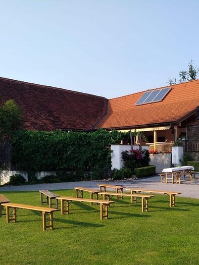 Eine ruhige Grünfläche mit Holzbänken und Tischen. Im Hintergrund ist ein traditionelles Gebäude mit einem roten Dach und Blumenschmuck zu sehen. | © Buschenschank Wiesenhofer