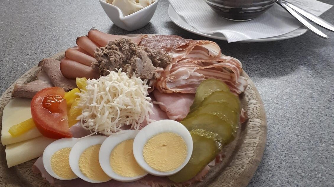 Eine appetitliche Platte mit verschiedenen Wurst- und Käsevariationen, Eiern und Gemüse. Im Hintergrund ein Dessert aus Schokolade und Obst. | © Buschenschank Wiesenhofer