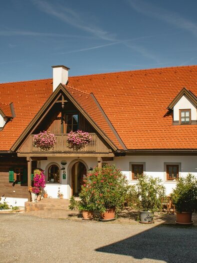 Ein traditionelles deutsches Haus mit einem roten Zeltdach und grünen Fensterläden. Der Eingangsbereich ist von Blumen und Pflanzen umgeben. | © Weinbauernhof Wildbacher | Fam. Wildbacher