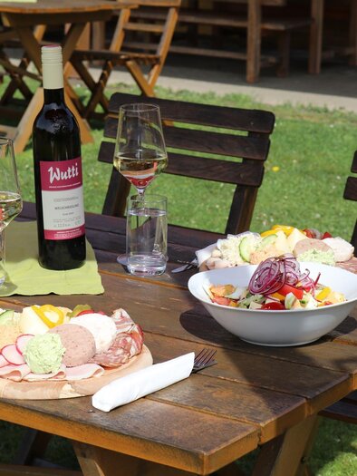 A pretty table outdoors with a selection of sausage and cheese platters, along with a colorful salad. On the table, there is also a bottle of wine and glasses. | © Andreas Wutti | Weingut Buschenschank Wutti