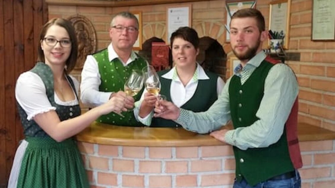 Eine Gruppe von vier Personen in traditionellen Trachten steht an einer Bar. Sie stoßen mit Gläsern an und lächeln freundlich. | © Andreas Wutti | Weingut Buschenschank Wutti